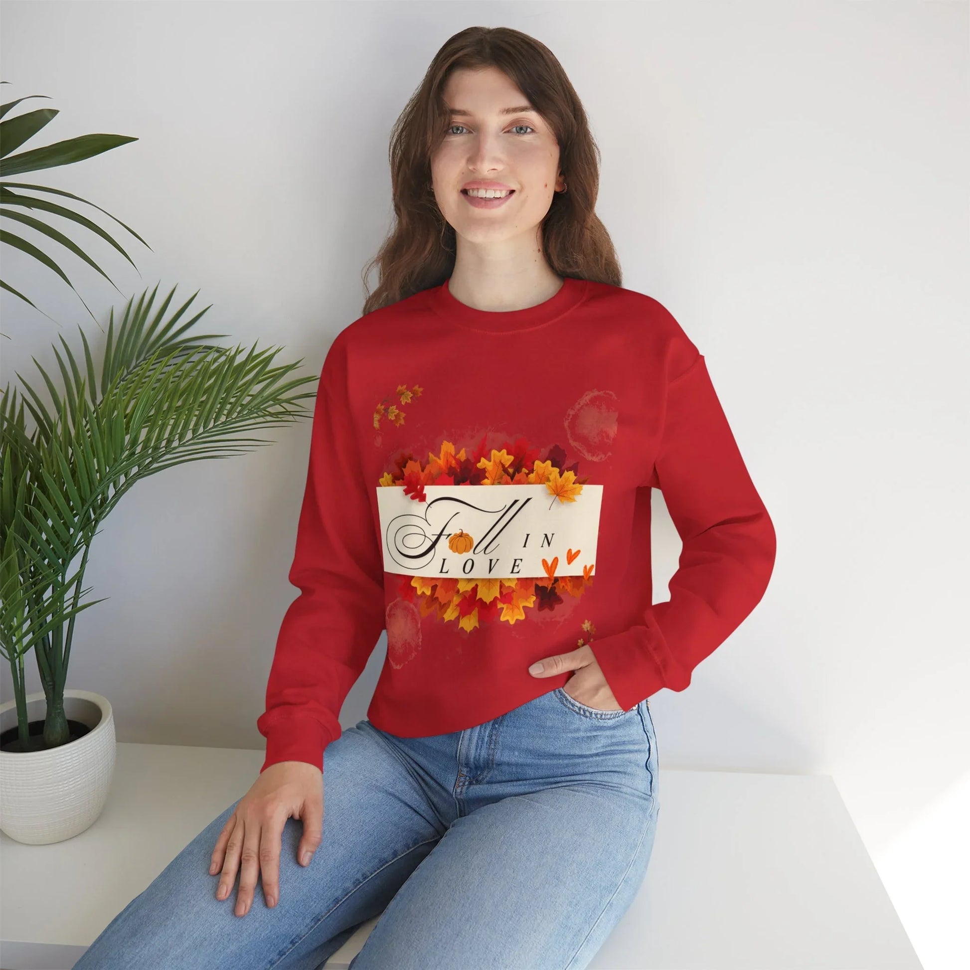 Woman in red Fall in Love sweatshirt with autumn leaves graphic, sitting by potted plant